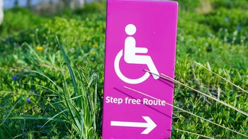 Foto von Yomex Owo auf Unsplash Rosa und weißer Hinweis mit Behindertenlogo. Pfeil zeigt nach rechts mit Rasen im Hintergrund