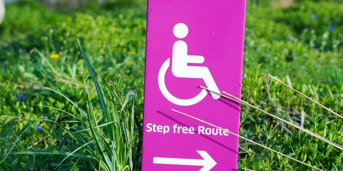 Rosa und weißer Hinweis mit Behindertenlogo. Pfeil zeigt nach rechts mit Rasen im Hintergrund