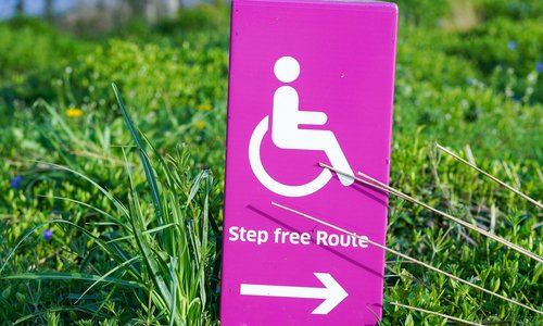 Rosa und weißer Hinweis mit Behindertenlogo. Pfeil zeigt nach rechts mit Rasen im Hintergrund